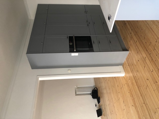 Kuvassa on harmaa keittiökaappi, jossa on uuni, valkoiset seinät ja puulattia. Huoneessa on myös valkoinen ovi ja musta laukku lattialla.