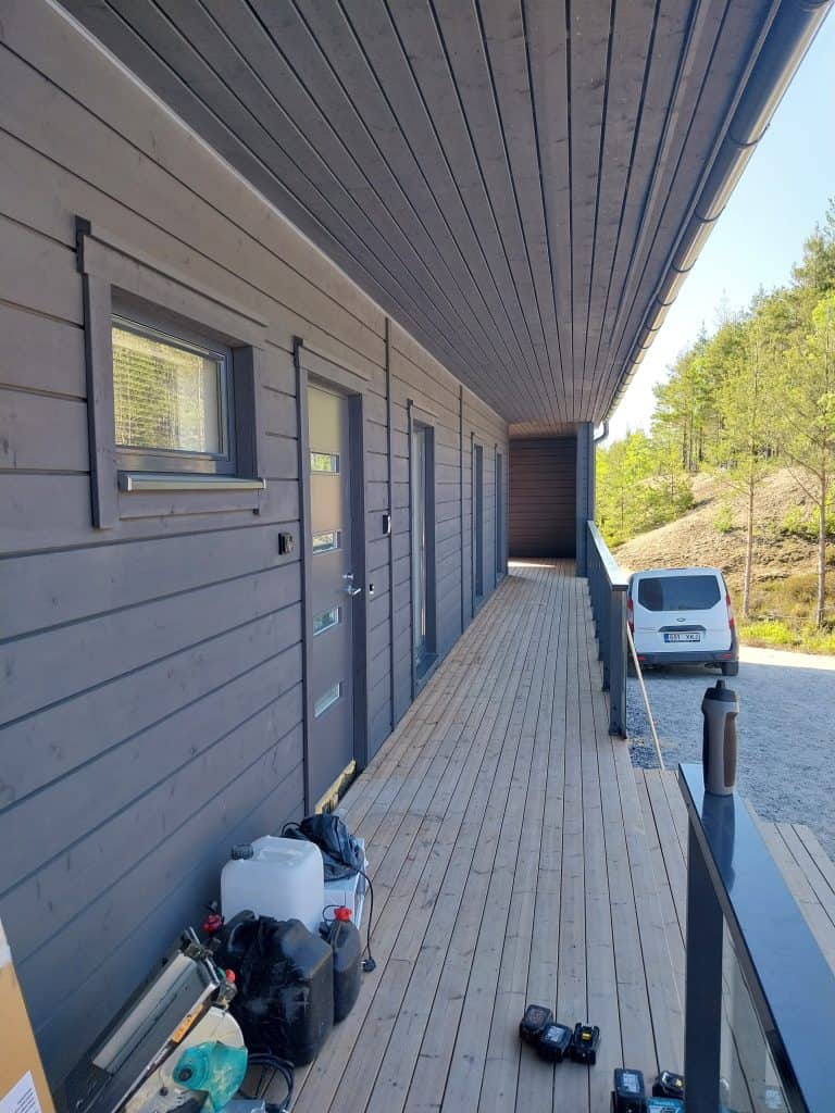 Ulkokuva rakennuksesta, jossa on harmaa puuverhous, ikkunoita, ovia ja katettu puinen kävelytie, jonka päässä näkyy valkoinen pakettiauto.