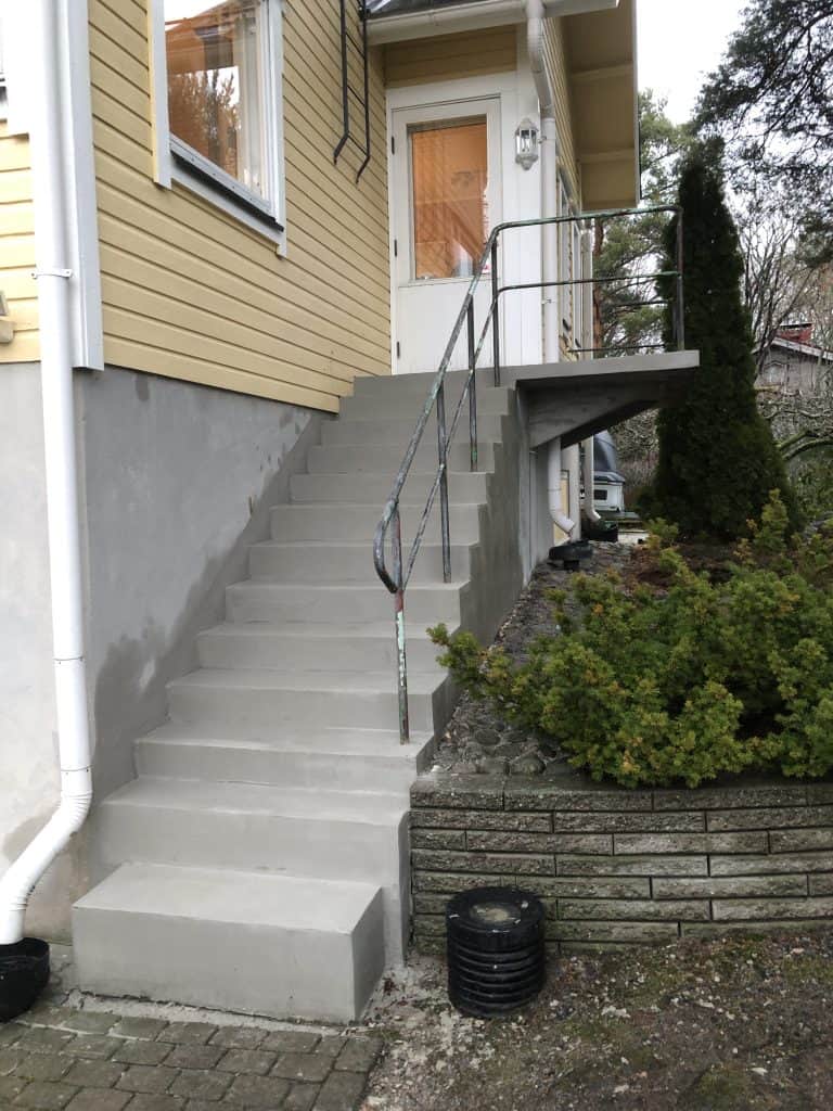 Ulkona on keltaisen talon harmaat portaat, joissa on metallinen kaide ja vihreitä pensaita.