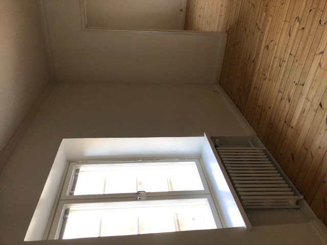 Huoneessa on puulattia, valkoinen seinä ja ikkuna, jonka alla on valkoinen patteri.