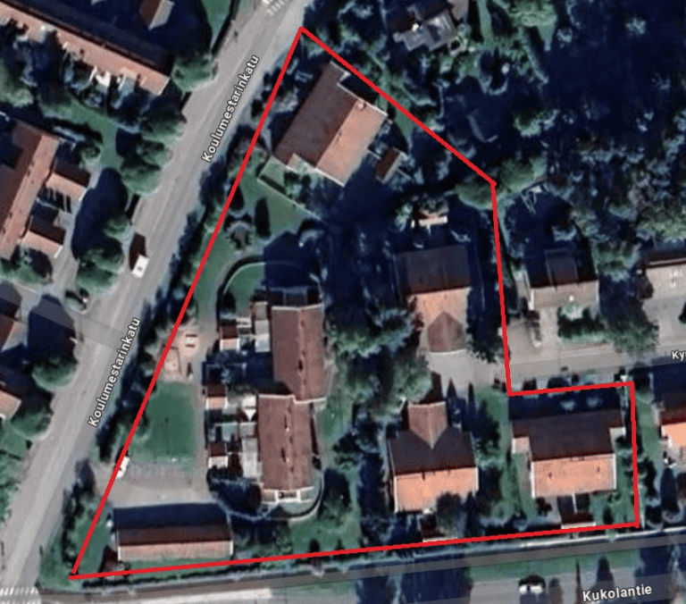 Ilmakuva alueesta, jota rajaa punainen viiva, jossa on rakennuksia, puita ja katuja aurinkoisessa säässä.