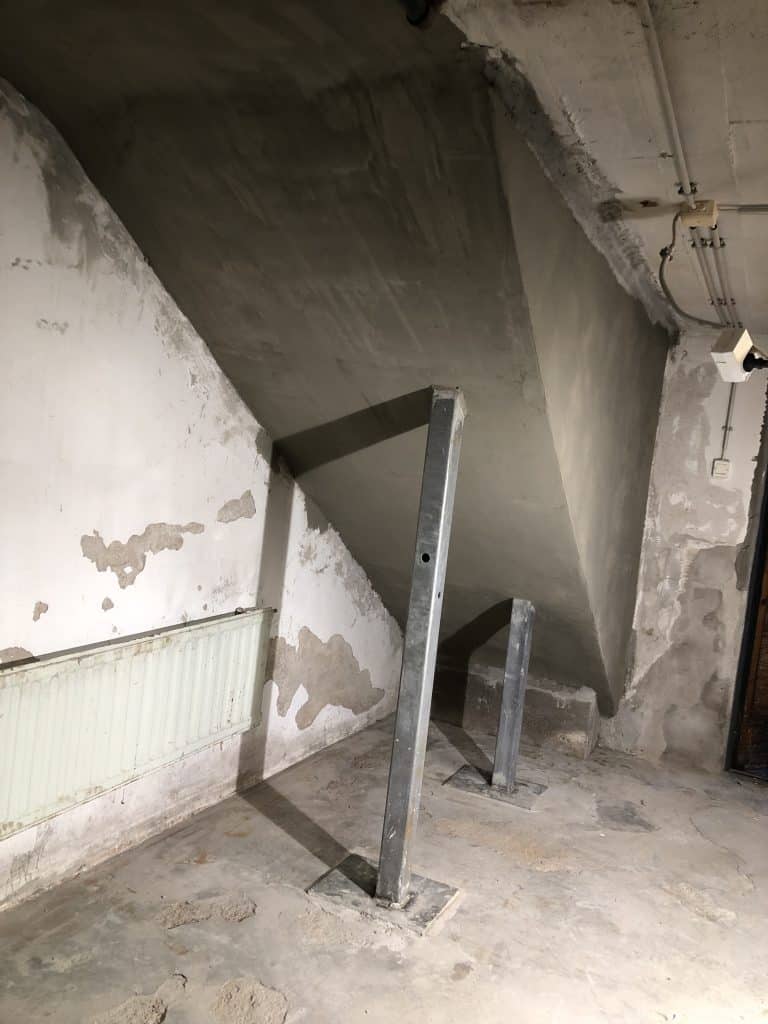 Kellaritila, jossa on betoninen katto, tukipylväät, patteri ja rapistuva seinä, valaistus on himmeää.