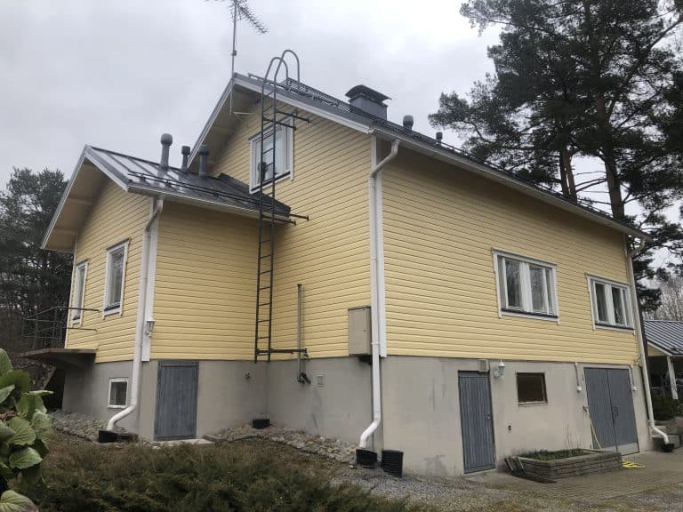 Kaksikerroksinen, keltainen puutalo, jossa on harmaa kivijalka, musta katto ja musta tikkaat.