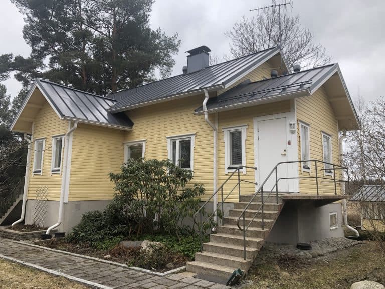 Kaksikerroksinen keltainen puutalo, jossa on harmaa katto, valkoiset ikkunat ja portaat, jotka johtavat valkoiselle ovelle.