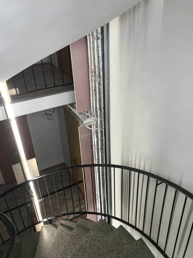 Ylhäältä kuvattu kierreporras, jossa on mustat kaiteet ja harmaat kiviset portaat, sekä putkia ja ovia taustalla.