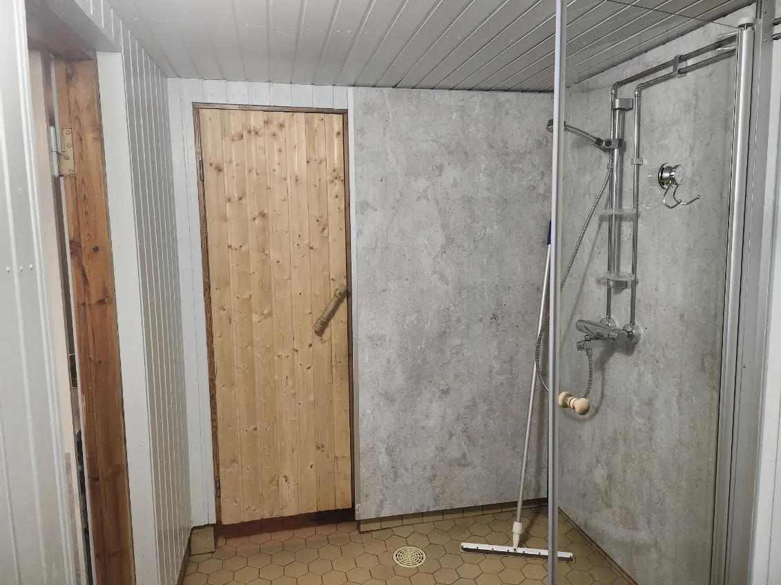 Kylpyhuoneen sisätila, jossa on puinen ovi, harmaa seinäpaneeli ja suihkukaappi, lattia on kuusikulmainen laatta.