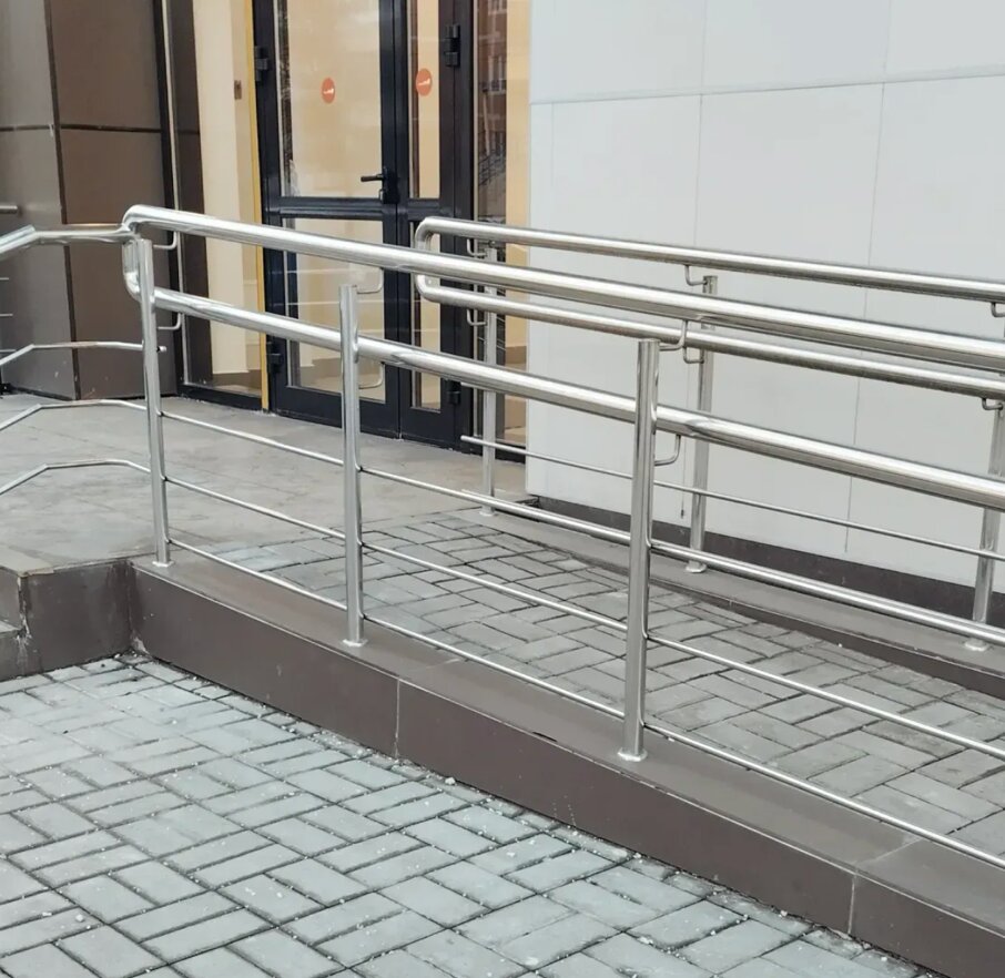 Kuva metallisesta luiskan kaiteesta, jossa on harmaat kivilaatat ja ruskea pohja, kirkkaassa päivänvalossa.