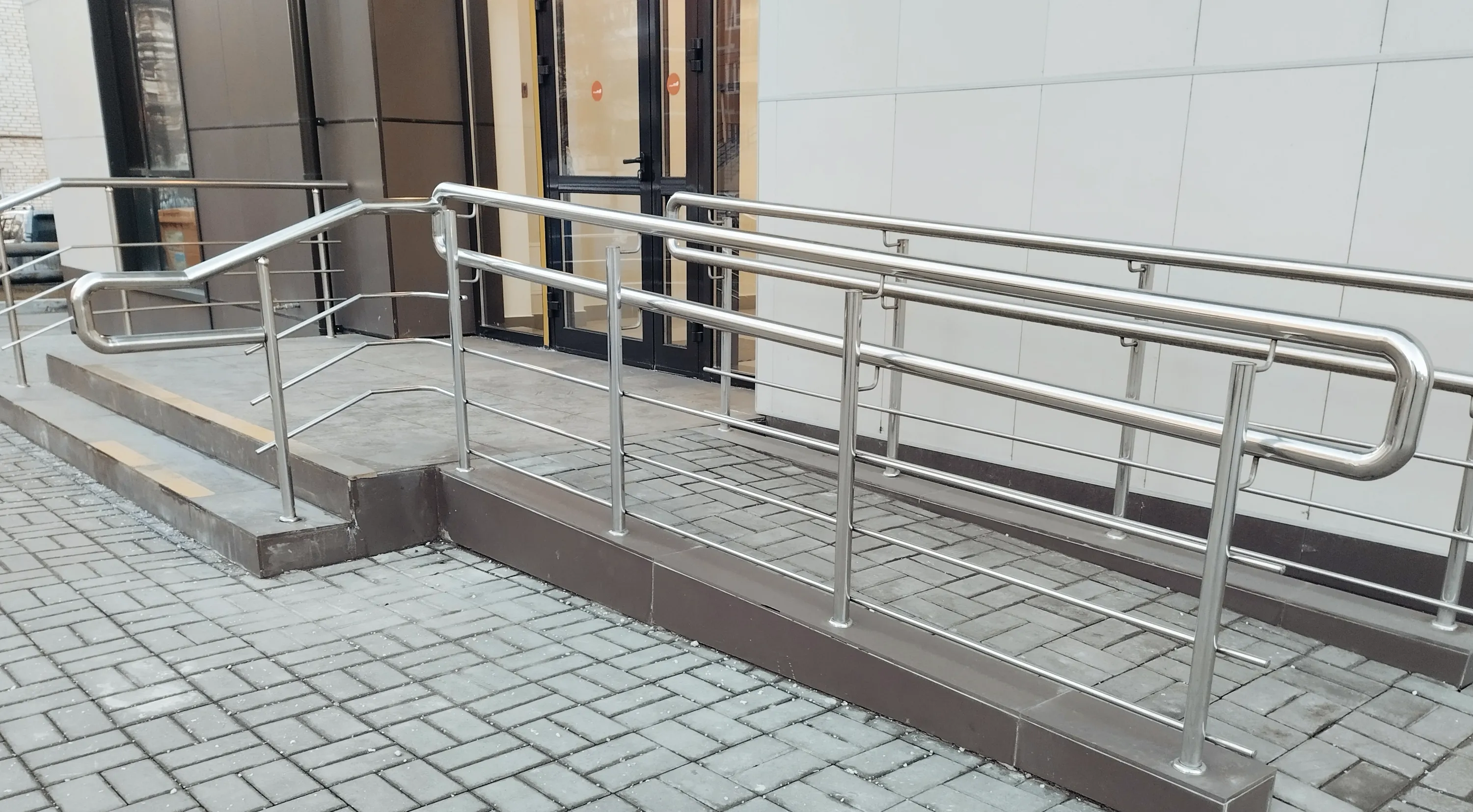 Kuva näyttää ulkona olevan luiskan ja portaat, joissa on metalliset kaiteet. Luiska ja portaat johtavat rakennuksen sisäänkäynnille, jossa on lasiovet. Maassa on harmaita kivilaattoja.