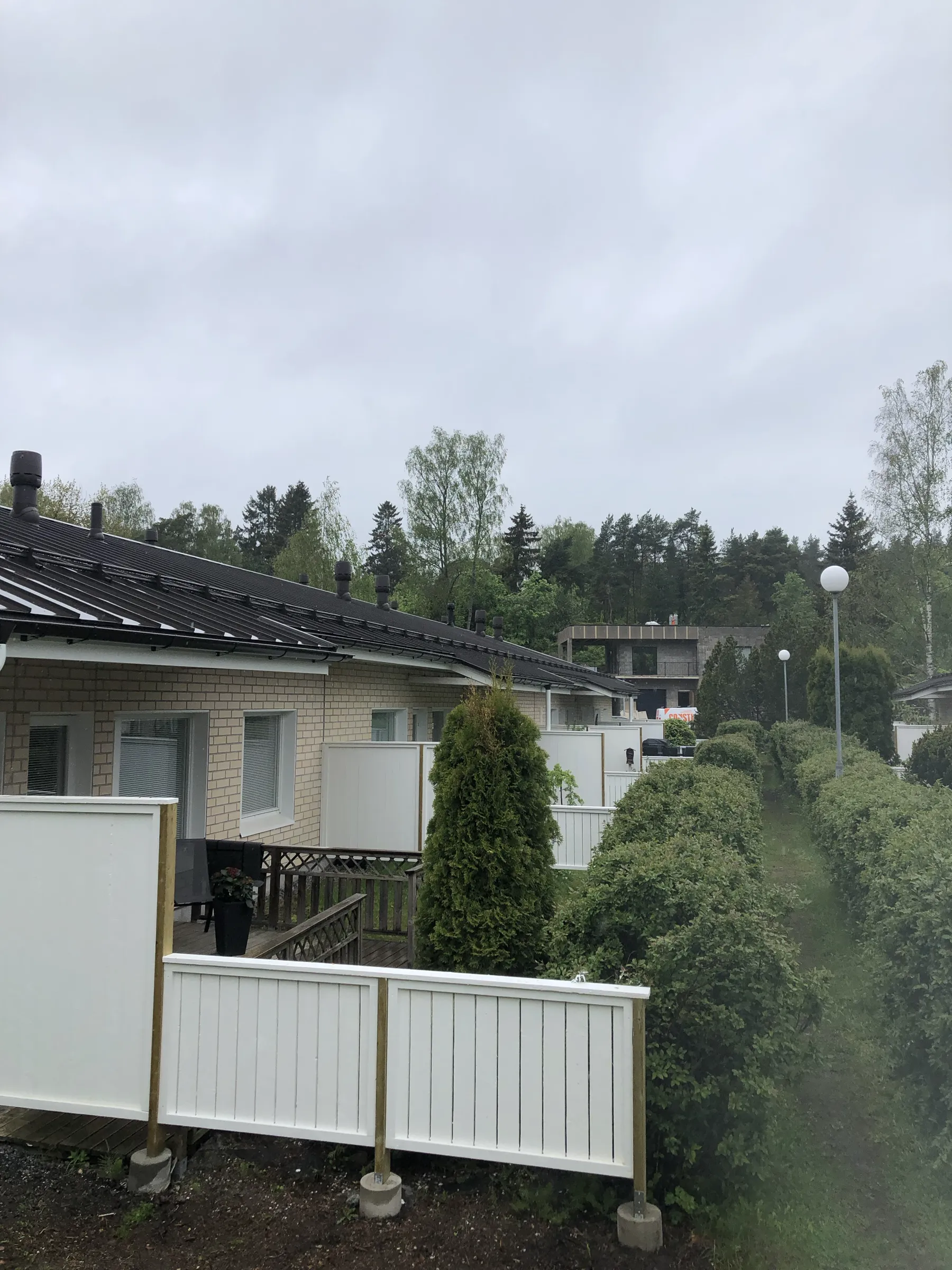 Matala tiilitalo, valkoiset aidat, vihreät pensaat ja harmaa taivas.