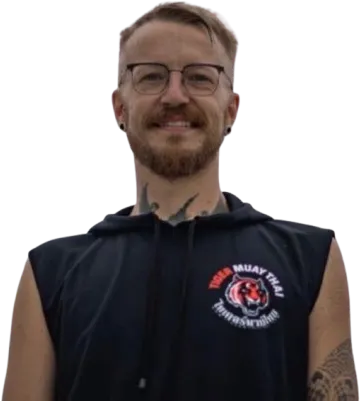 Hymyilevä mies silmälaseissa, parrassa ja tatuoinneissa, yllään hihaton huppari, jossa on Tiger Muay Thai -logo, mustalla taustalla.