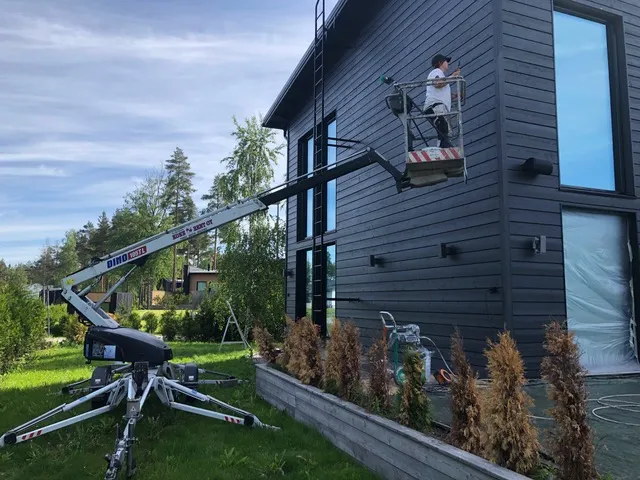 Ulkona on Dino-nosturi, jonka korissa seisoo henkilö maalaamassa tummaa puutaloa. Nosturi on asetettu nurmikolle, ja talon edessä on pieniä pensaita.