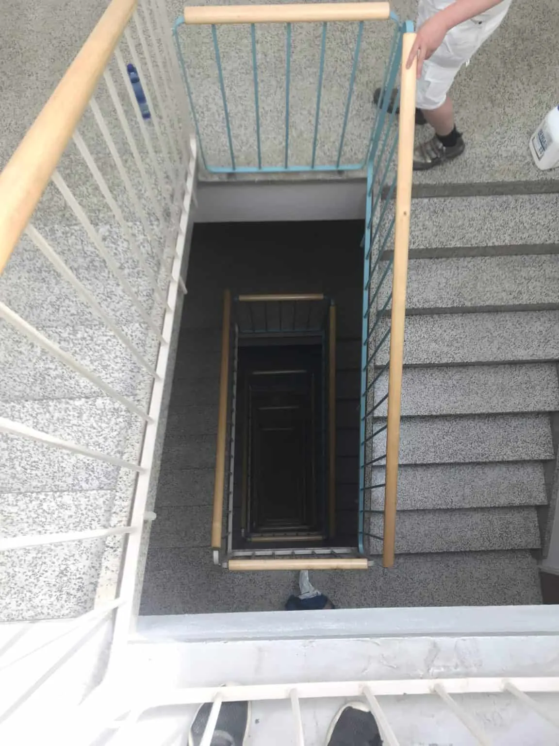 Ylhäältä kuvattu näkymä portaikkoon, jossa on valkoiset ja vaaleansiniset kaiteet. Portaat ovat harmaat ja valaistus on heikko, syvällä portaikossa on pimeää.