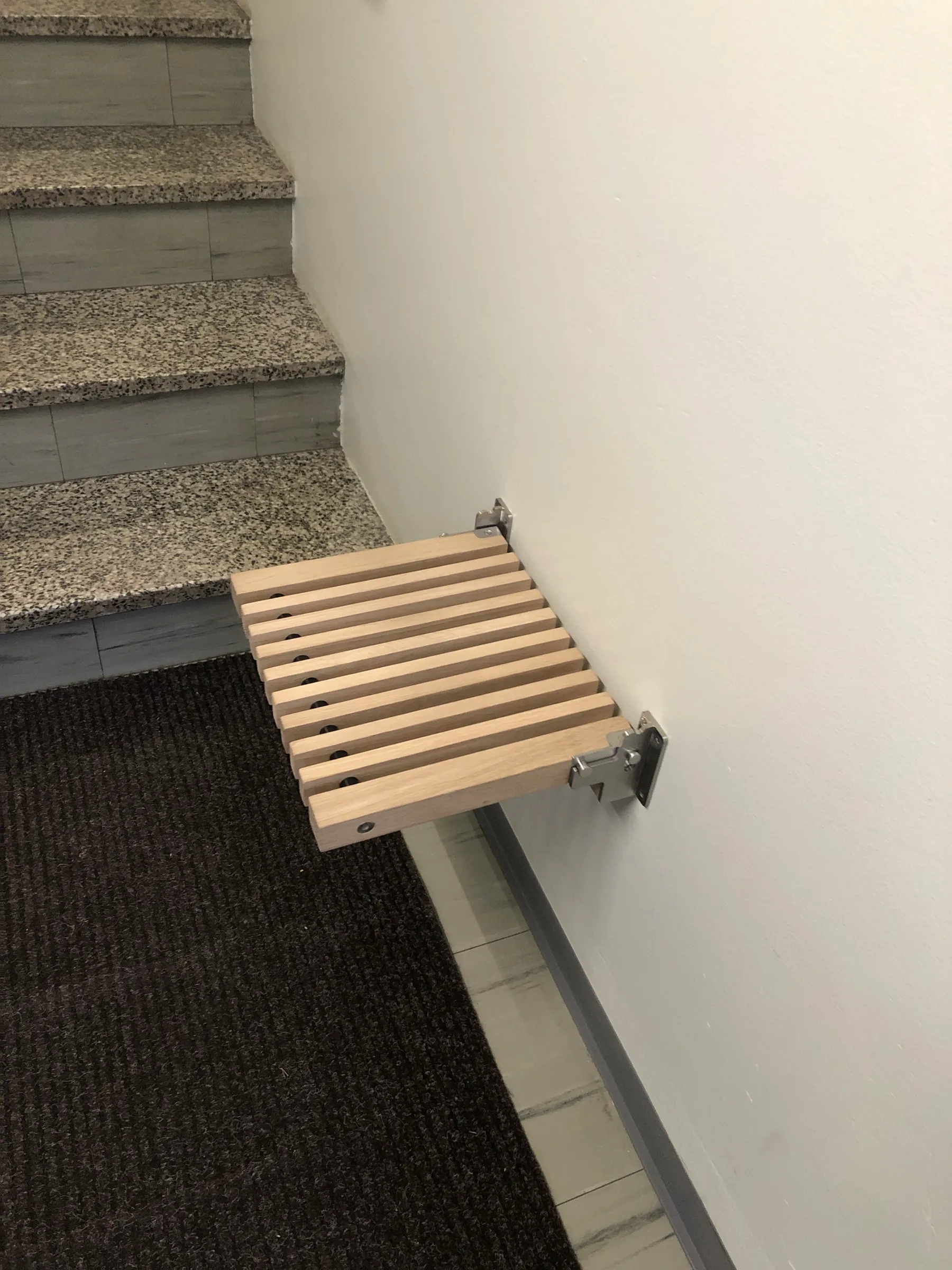 Seinään kiinnitetty puinen istuin, portaat ja tumma matto näkyvät. Istuin on valmistettu vaaleista puurimoista ja metallisista kiinnikkeistä.