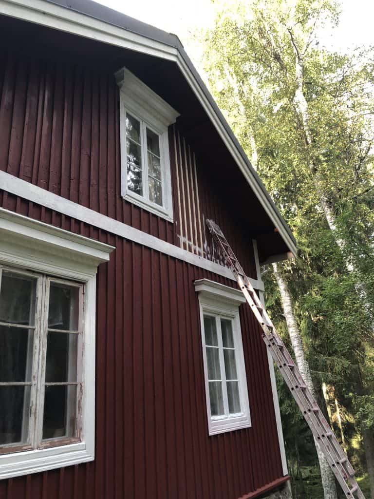 Punainen puutalo, jossa on valkoiset ikkunakehykset ja tikkaat nojaavat seinään, valoisassa ulkoilmassa.