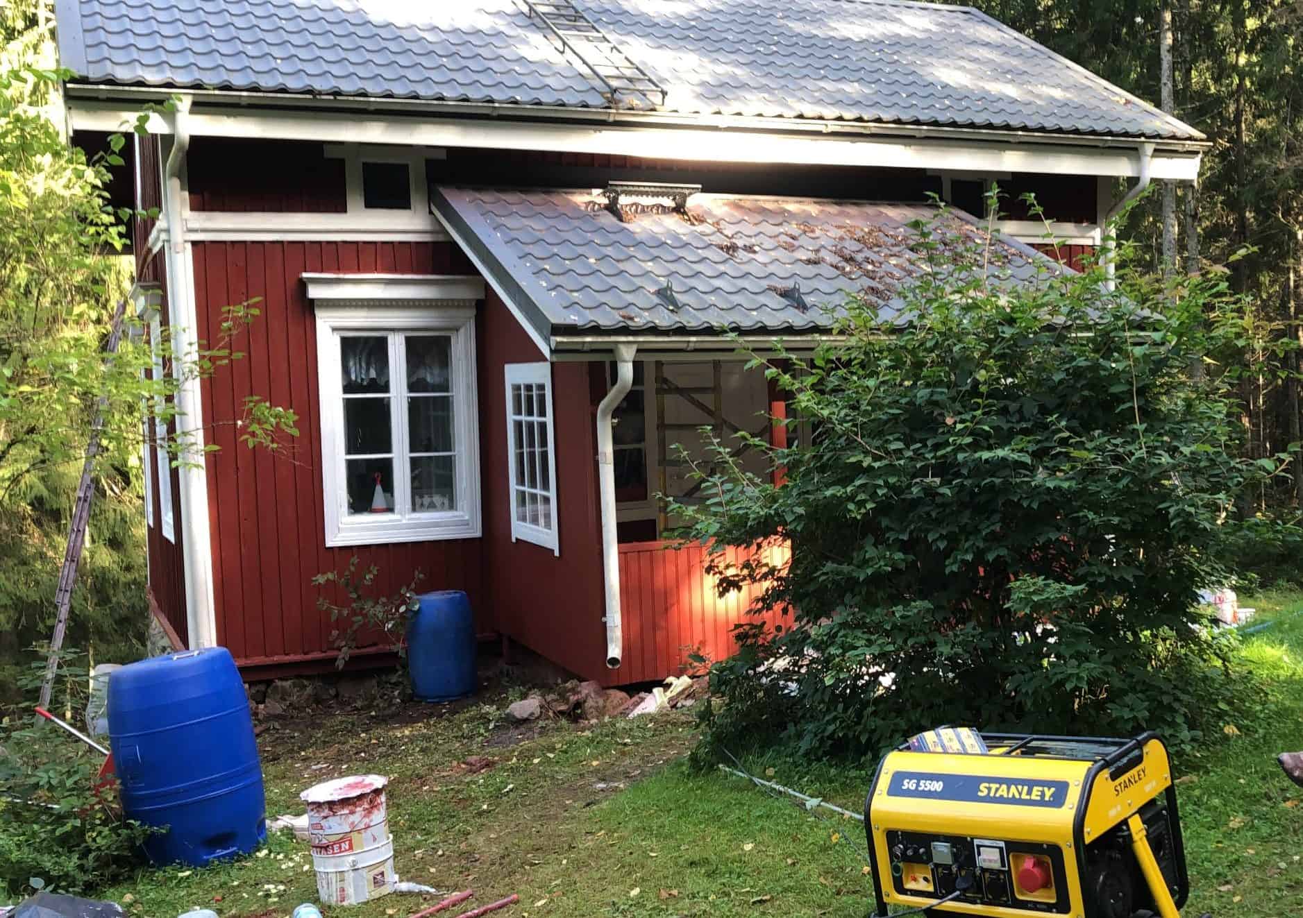 Punainen puutalo, jossa harmaa peltikatto, valkoiset ikkunankarmit ja räystäät, sinisiä tynnyreitä ja keltainen aggregaatti nurmikolla, valaistus on luonnollinen.