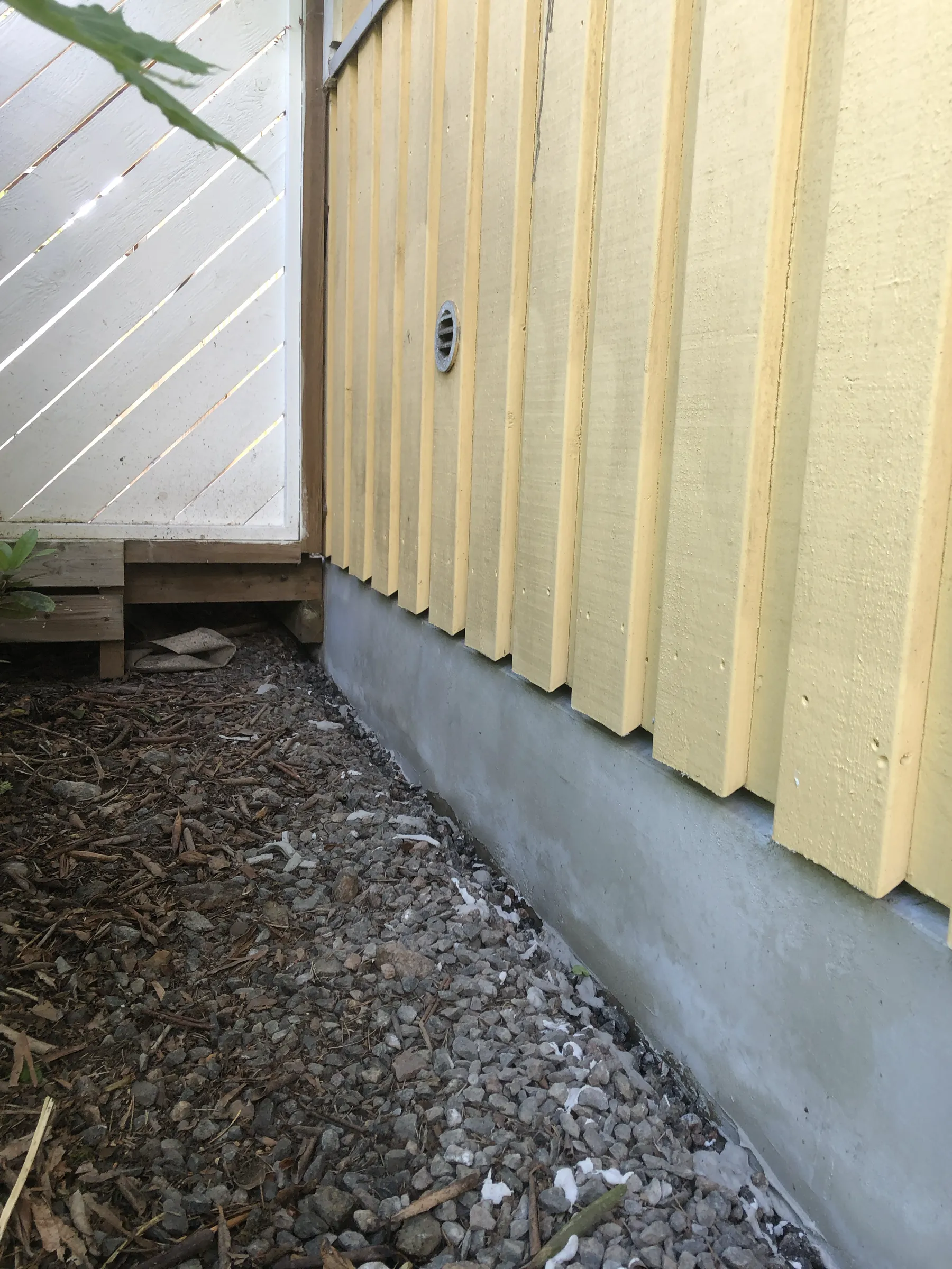 Kuva näyttää keltaisen puurakennuksen harmaalla betonijalustalla, jonka vieressä on valkoinen aita ja maassa on kiviä ja lehtiä. Valo on luonnollinen ja pehmeä.