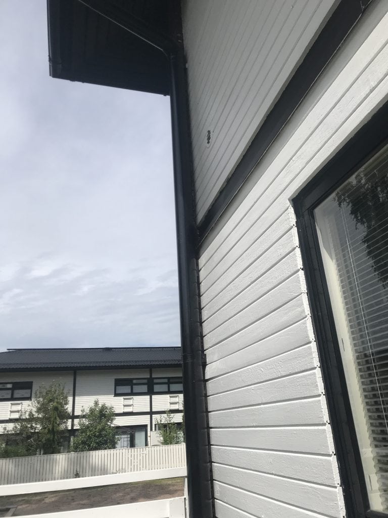 Ulkokuva valkoisesta puutalosta, jossa on mustat yksityiskohdat, kuten ikkunanpuitteet ja sadevesijärjestelmä. Taustalla näkyy toinen rakennus ja osittain pilvinen taivas.