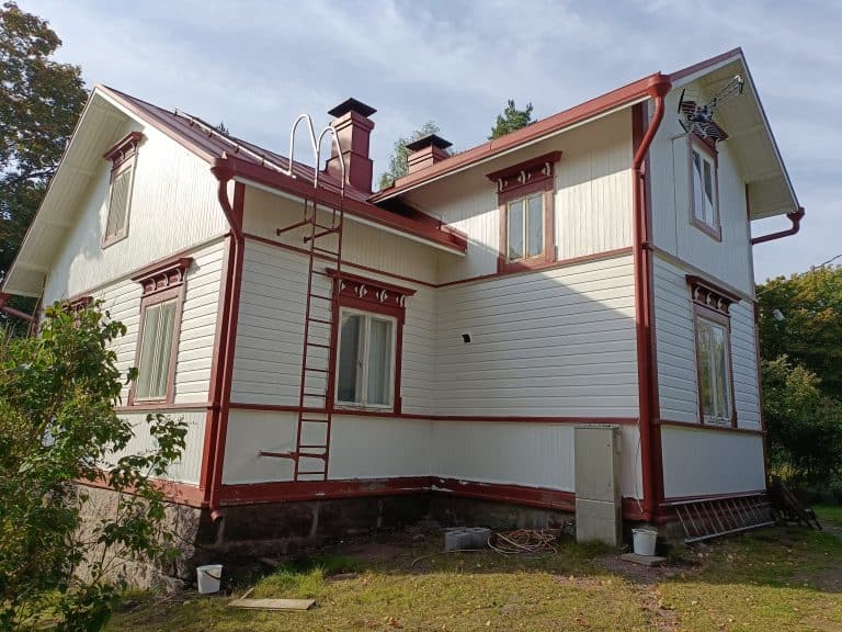 Kaksikerroksinen, valkoinen puutalo punaisilla koristeilla ja katolla, jossa on tikkaat ja savupiippu, aurinkoisessa säässä.