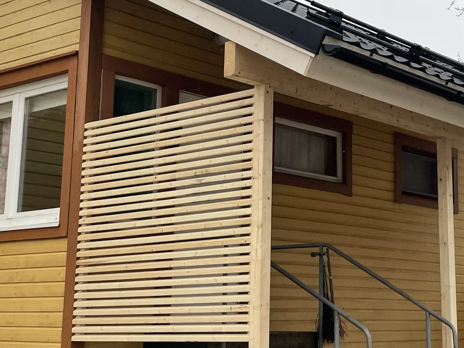 Vaaleankeltainen puutalo, jossa on puinen säleikkö ja harmaa metallinen portaikko.