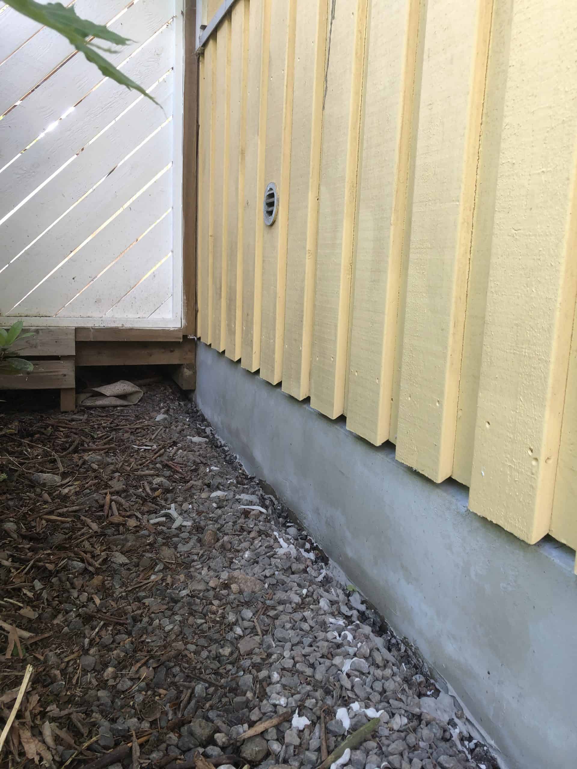 Ulkokuva keltaisesta puutalosta, jossa on harmaa kivijalka ja valkoinen puuaita vasemmalla, maassa on kuivia lehtiä ja kiviä.