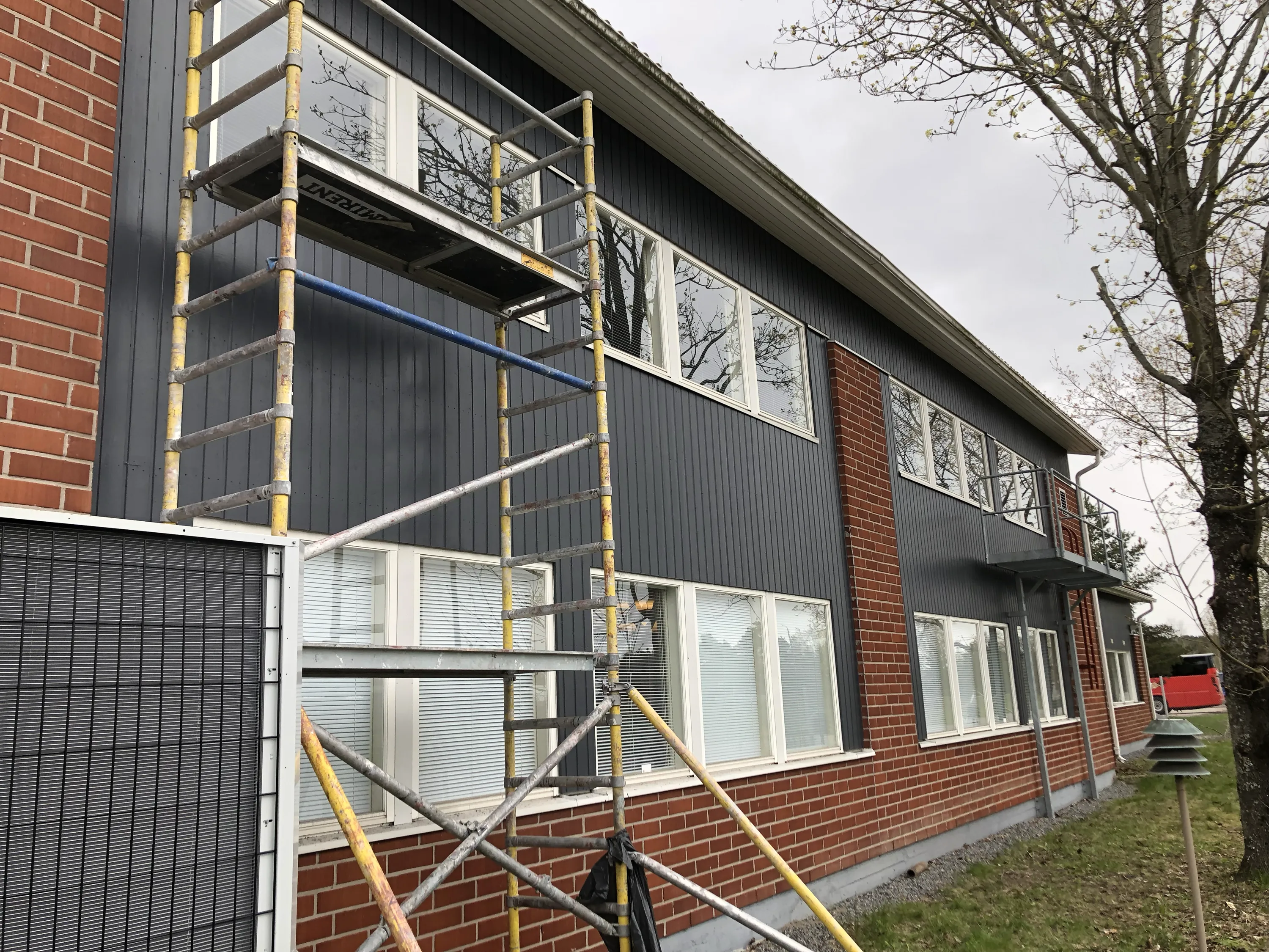 Rakennuksen ulkoseinää peittää teline, jossa on harmaa puuverhoilu, punatiilinen alaosa ja valkoiset ikkunat. Taustalla näkyy puu ja pilvinen taivas.