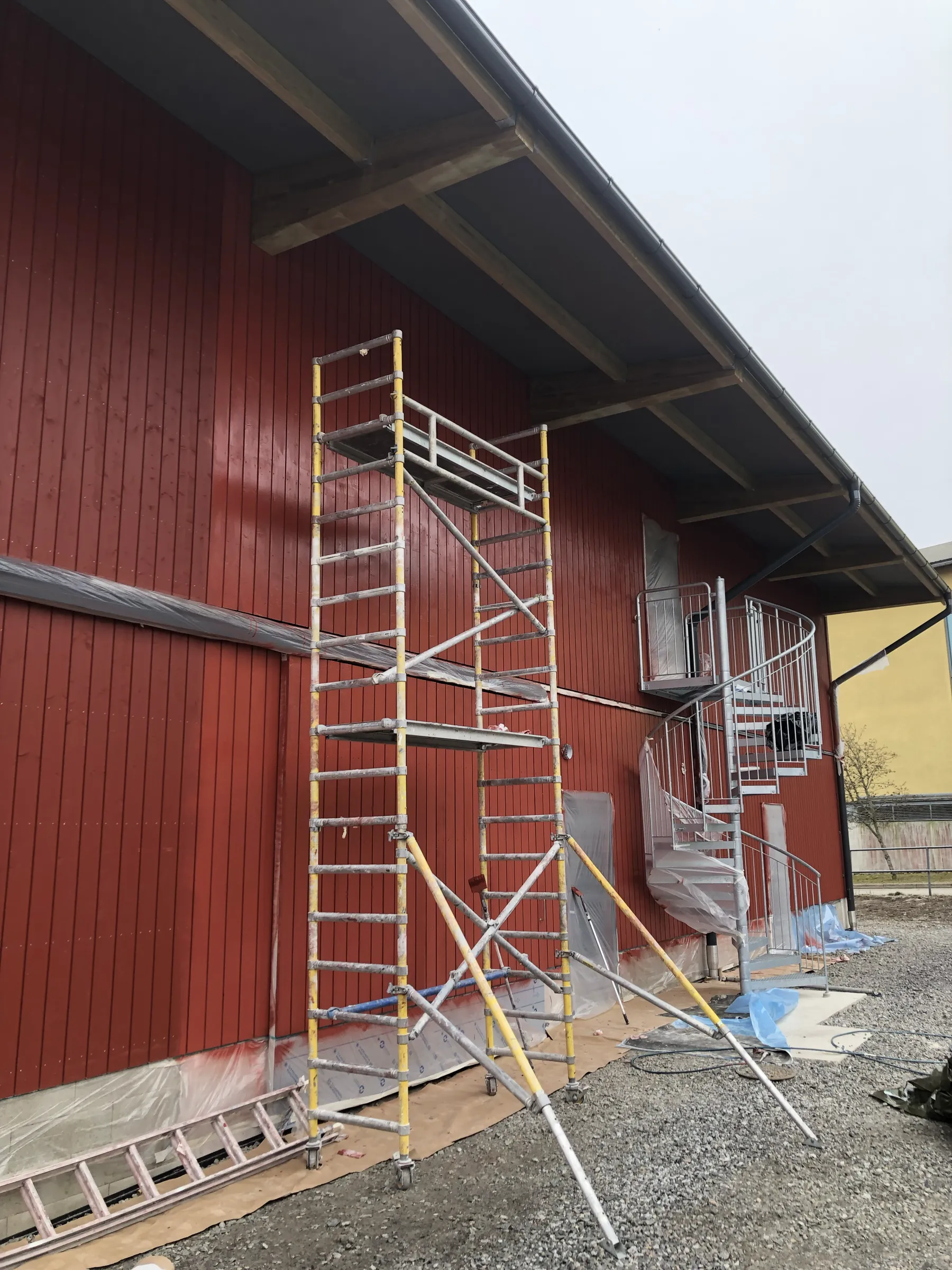 Ulkona on punainen puurakennus, jonka edessä on telineet ja kierreportaat. Rakennuksen katto on tumma ja taivas on pilvinen.