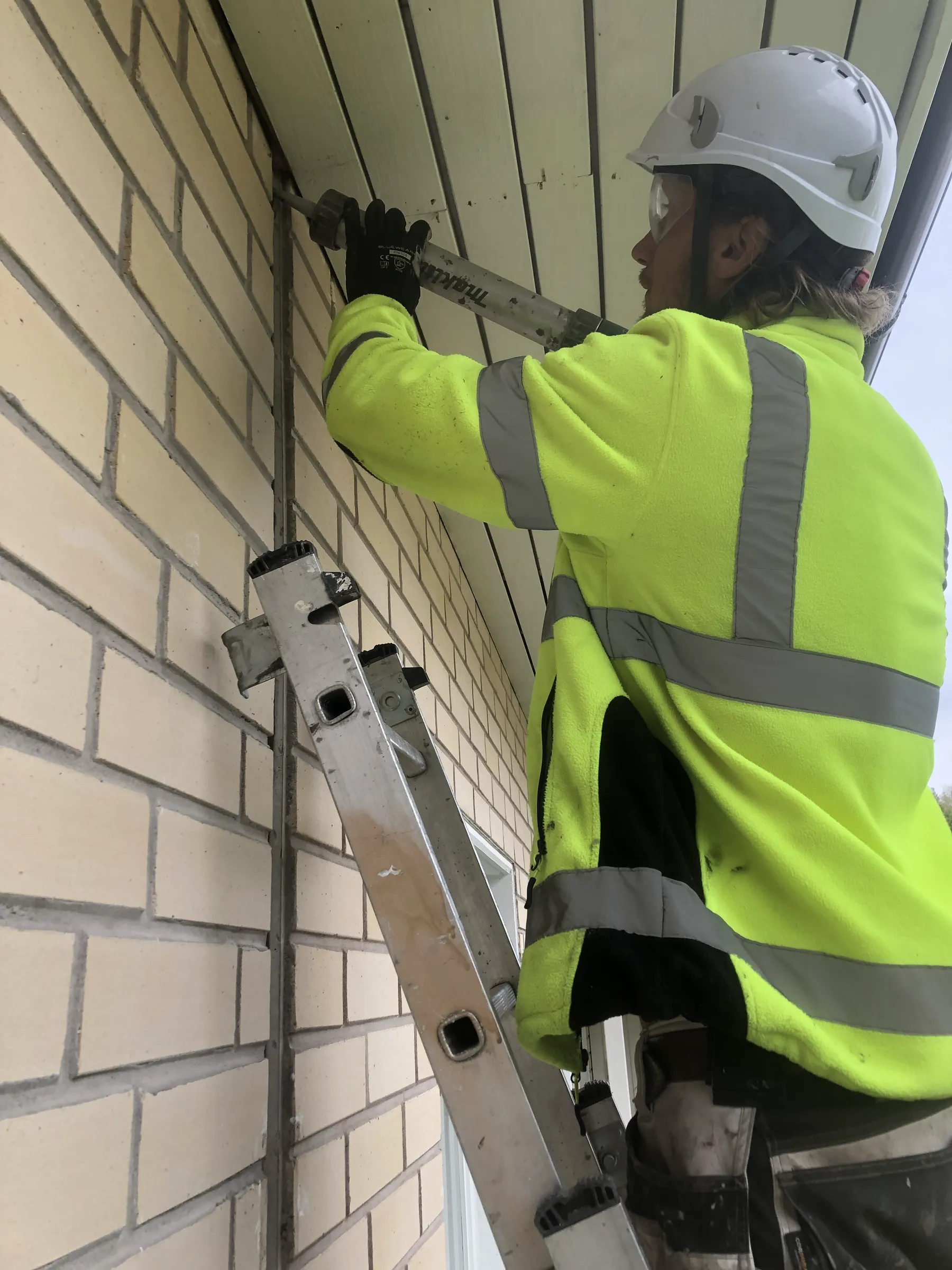 Rakennustyöntekijä tikkailla tiiliseinän vieressä, käyttäen tiivistyspistoolia, yllään suojakypärä, suojalasit ja huomiovärinen takki. Kirkas valaistus.