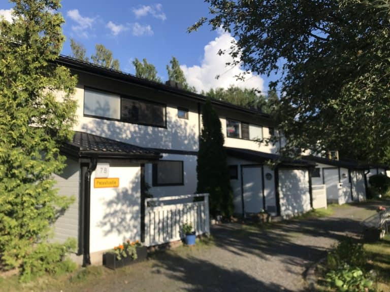 Rivitalo aurinkoisena päivänä, jossa on valkoiset seinät, mustat katot ja vihreät puut.