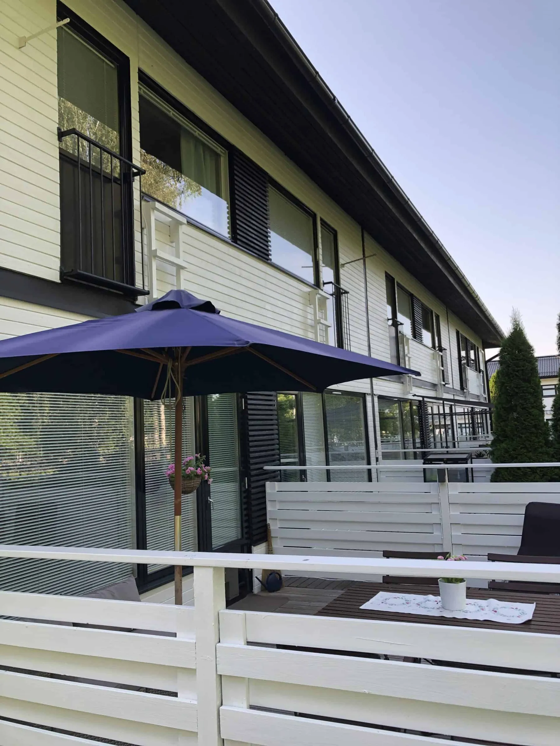 Ulkonäkymä rivitalosta, jossa on valkoiset seinät, mustat ikkunankarmit ja parvekkeet. Etualalla on sininen aurinkovarjo ja valkoinen puuaita.