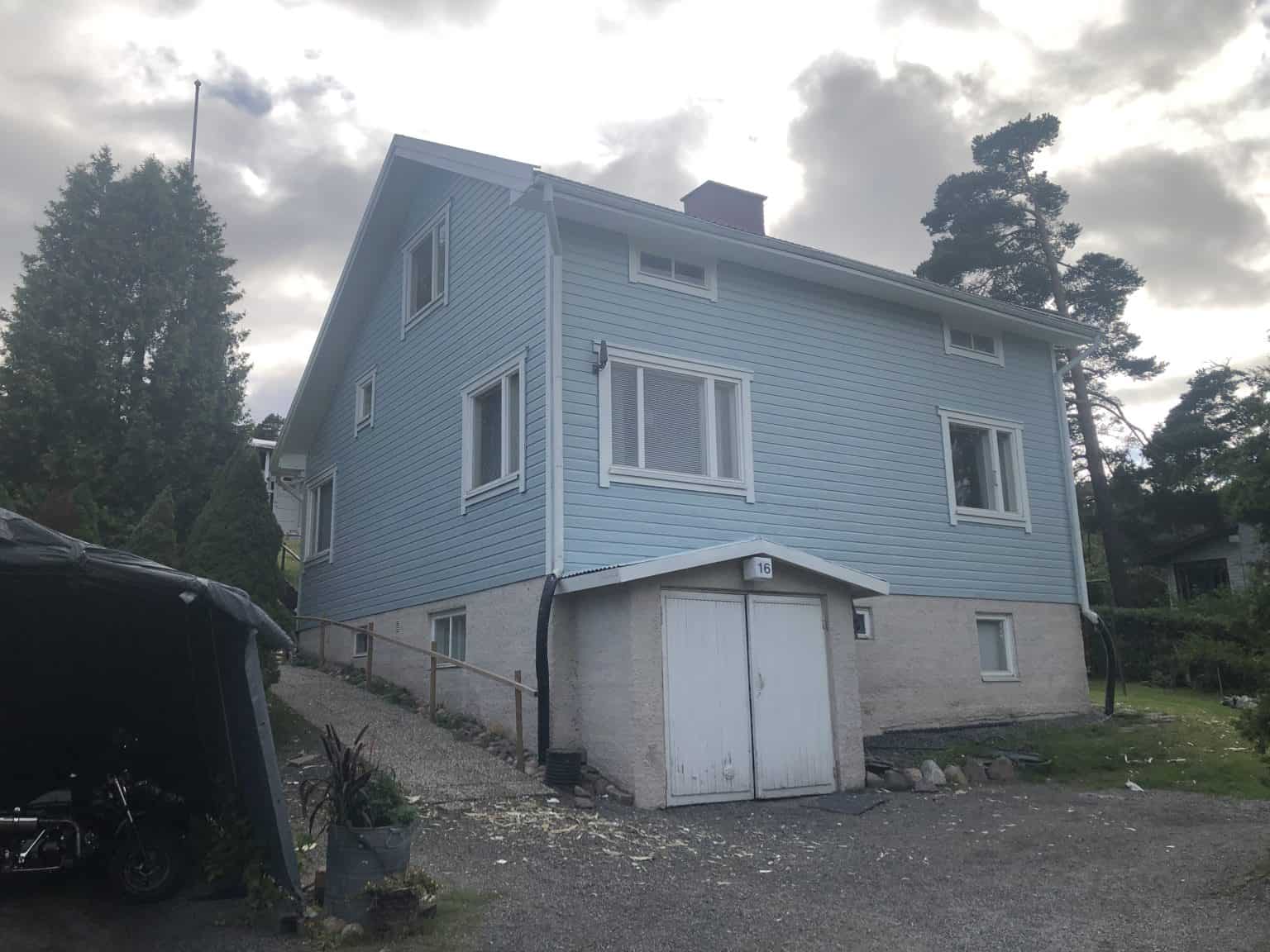 Kaksikerroksinen, vaaleansininen puutalo, jossa valkoiset ikkunat ja harmaa kivijalka, sijaitsee pilvisen taivaan alla. Etualalla näkyy katos moottoripyörällä ja kivetty piha.