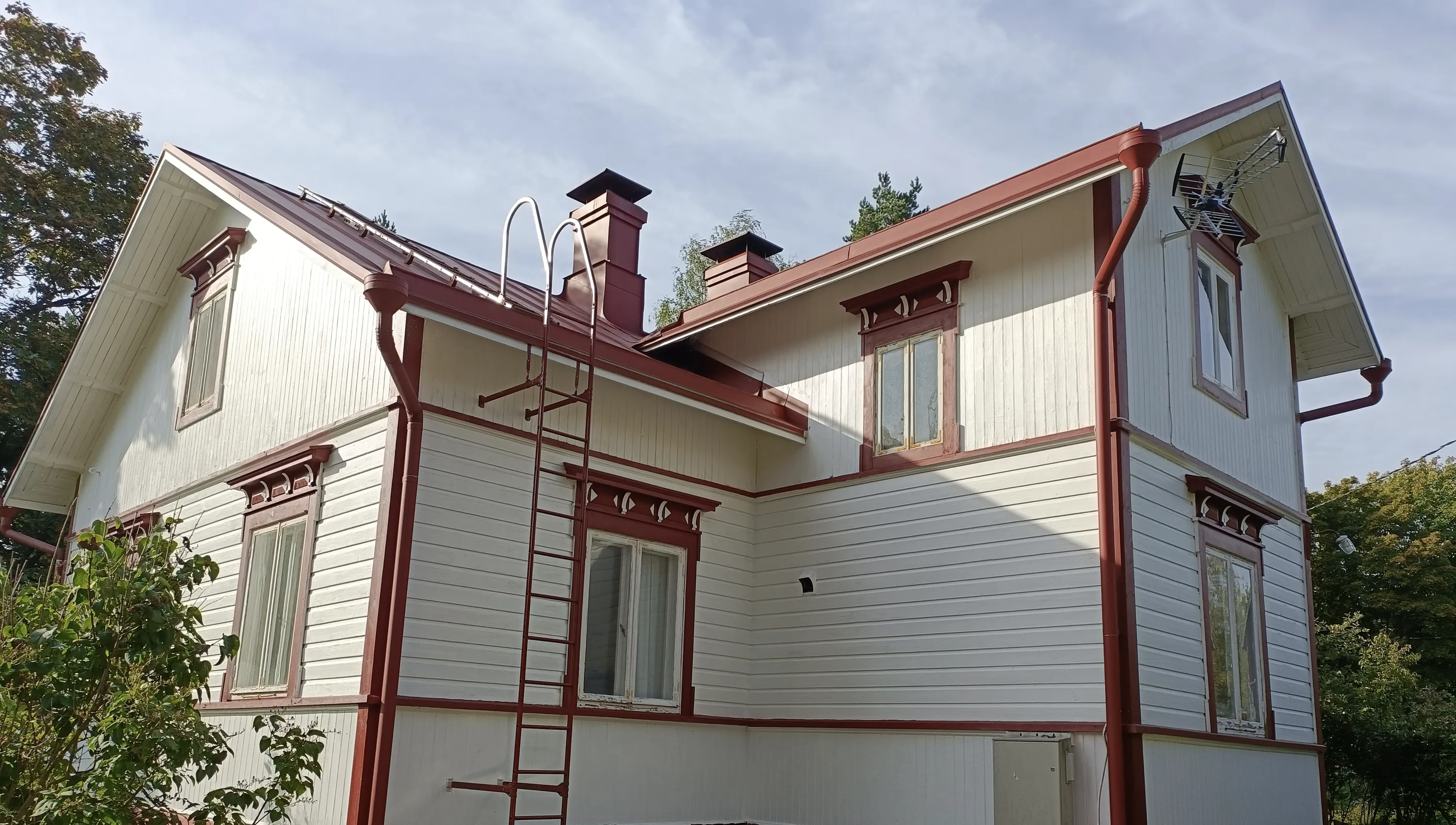 Vaalean talon julkisivu, jossa punaiset ikkunankehykset, katto ja rännit. Tikkaat johtavat katolle, jossa on kaksi savupiippua.