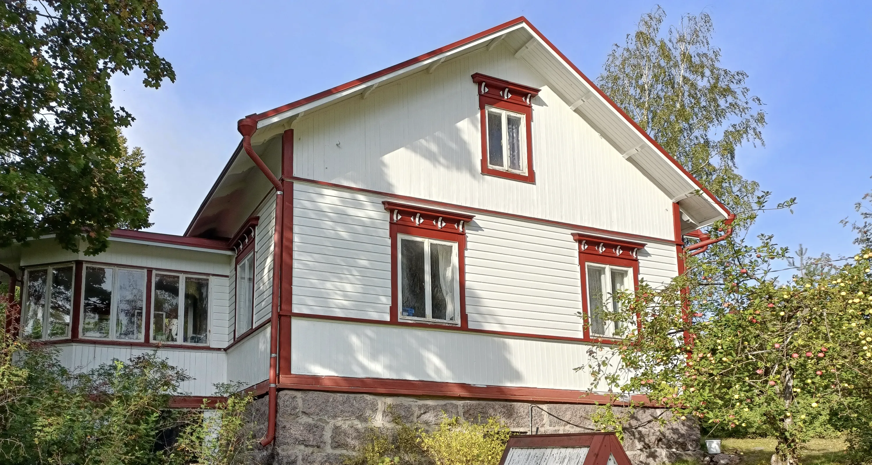 Valkoinen puutalo punaisilla yksityiskohdilla, kivijalka ja puutarha. Kirkas valo.