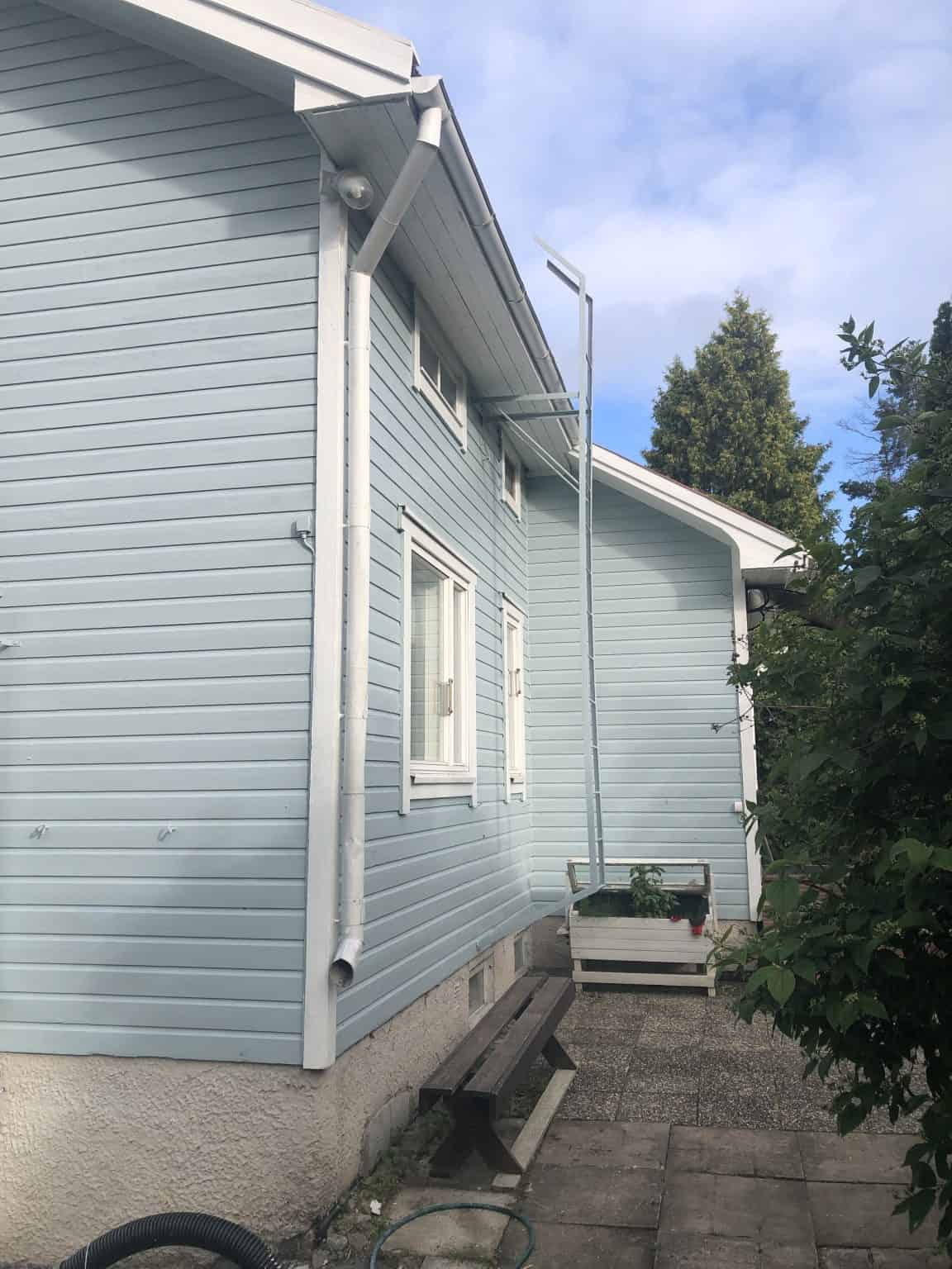 Vaaleansininen puutalo, valkoiset ikkunat ja syöksytorvi, puinen penkki ja kasvilaatikko. Kirkas päivä.