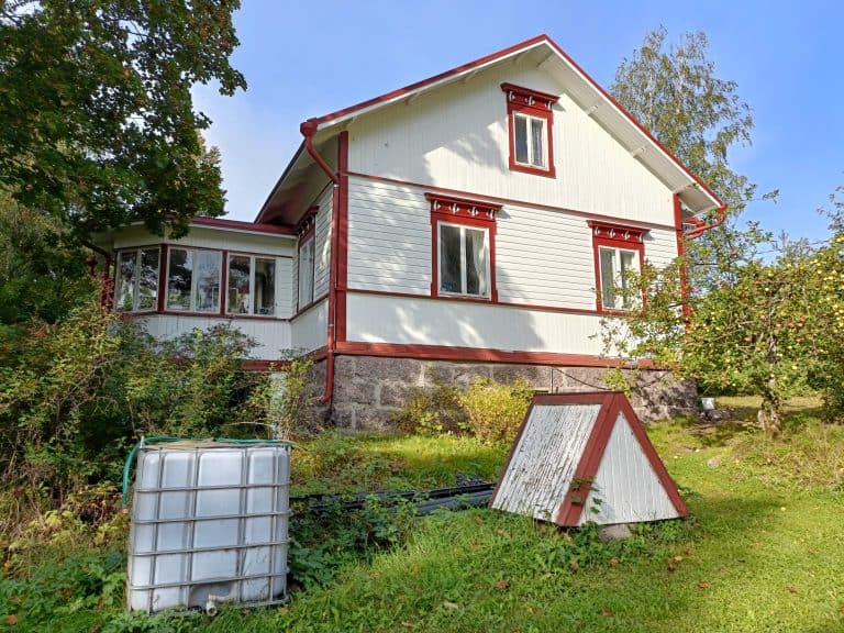 Kaksikerroksinen valkoinen talo punaisilla koristeilla, vihreä nurmikko ja puita aurinkoisena päivänä.