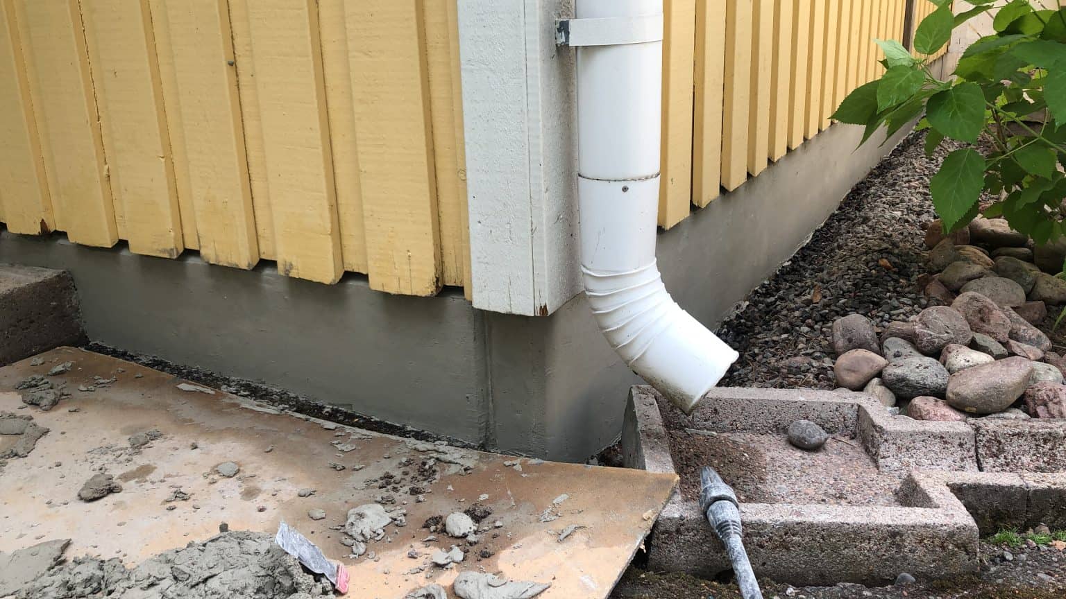 Kuva näyttää talon ulkokulman, jossa on keltainen puuverhous, harmaa betonijalka ja valkoinen syöksytorvi. Maassa on sementtijätettä ja kiviä.