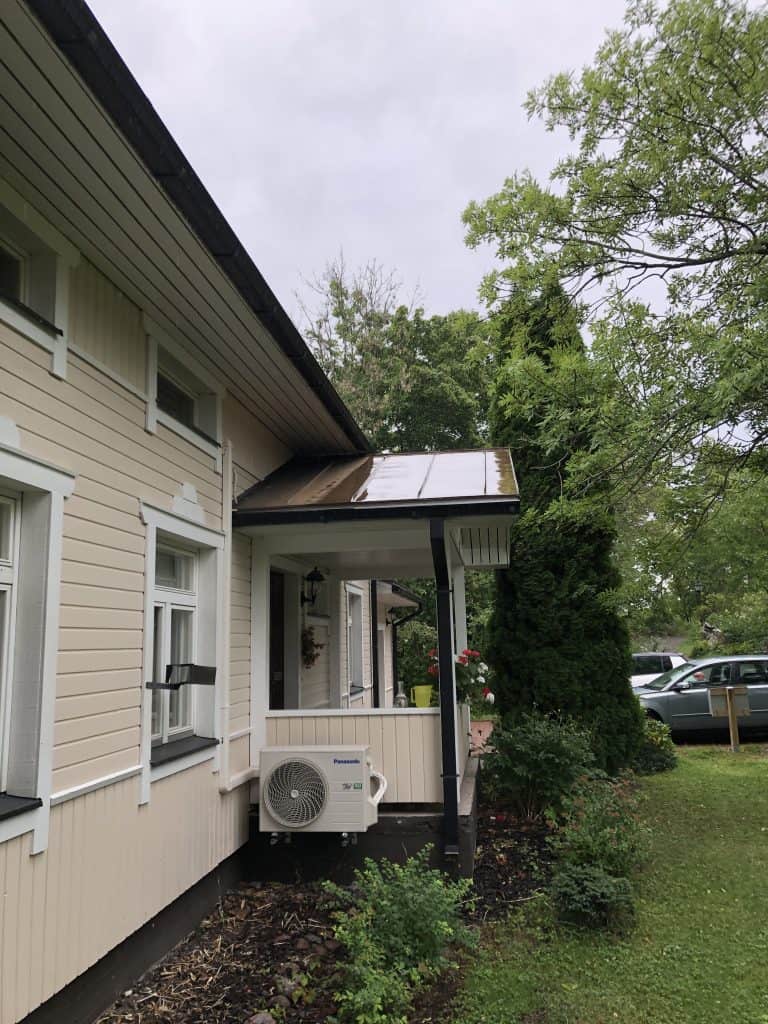 Vaalean talon ulkokuva, jossa on etukuisti, ilmastointilaite ja vihreä nurmikko. Taustalla näkyy puita ja autoja. Sää on pilvinen.