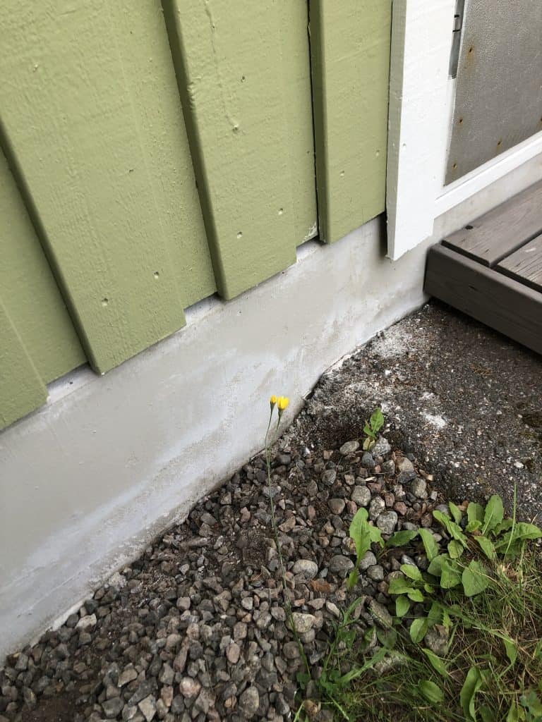 Lähikuva talon ulkoseinästä, jossa on vihreä puuverhous, harmaa kivijalka ja pieniä kiviä maassa. Kaksi keltaista kukkaa kasvaa kivien seassa.