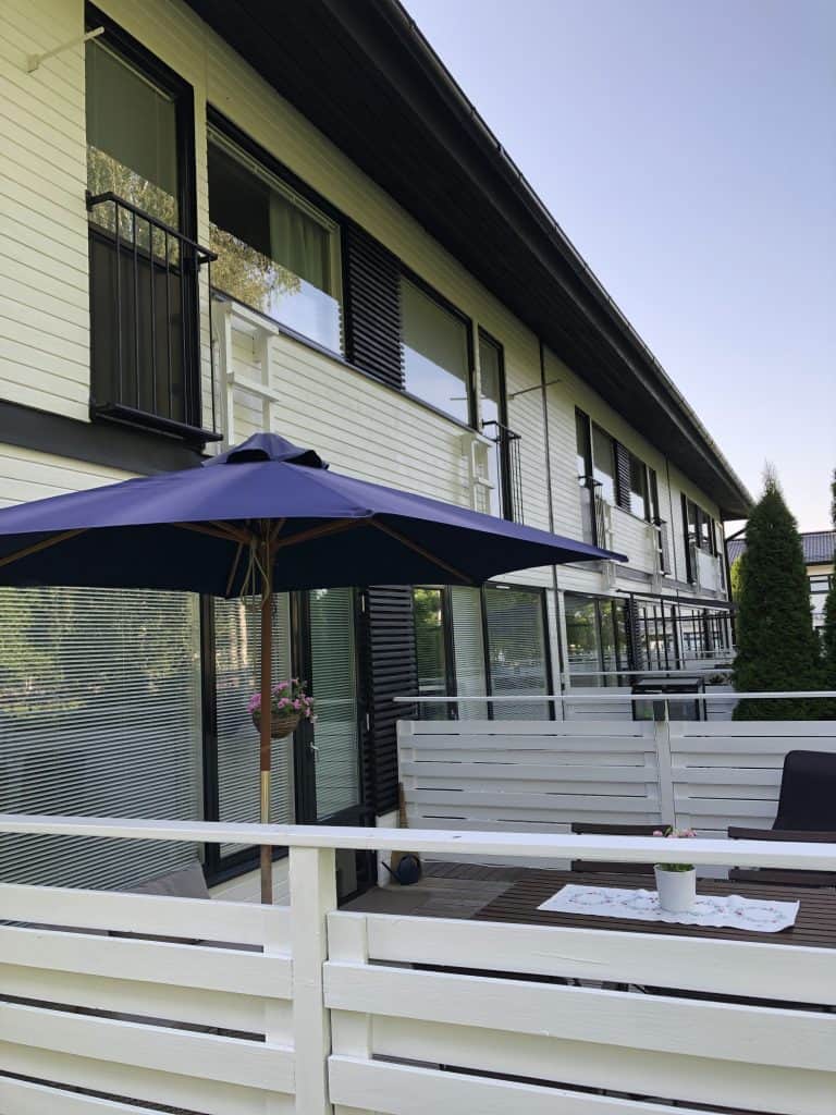 Ulkoterassi valkoisella aidalla, sininen aurinkovarjo, puinen pöytä ja tuoli, valoisa sää.