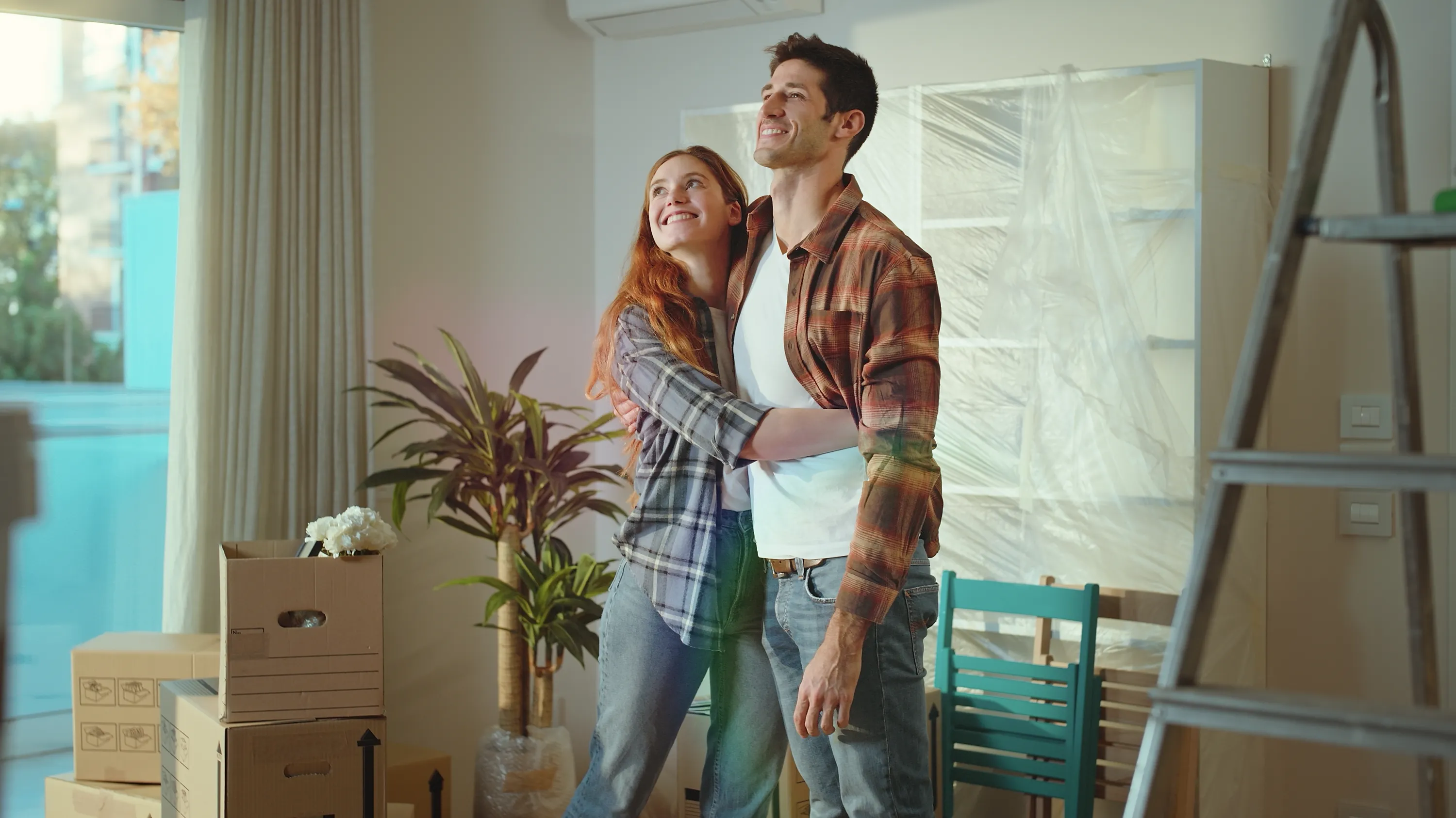 Nuori pariskunta halaa toisiaan uudessa asunnossa, jossa on muuttolaatikoita, kasveja ja maalaustarvikkeita. Nainen on punatukkainen ja pukeutunut ruutupaitaan, mies on ruskeahiuksinen ja hänellä on yllään ruutupaita ja valkoinen t-paita. Valoisa ja lämmin tunnelma.