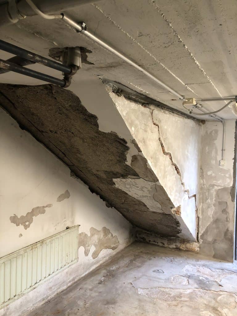 Huoneessa on vaurioitunut betonikatto, lohkeilevat seinät, putket katossa ja patteri seinällä, valaistus on himmeä.