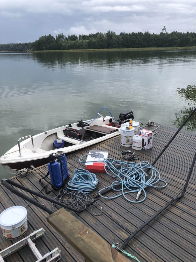 Kuvassa on vene laiturissa, jonka päällä on maalitölkkejä, johtoja ja muita tarvikkeita. Taustalla näkyy järvi ja metsäinen ranta pilvisen taivaan alla.